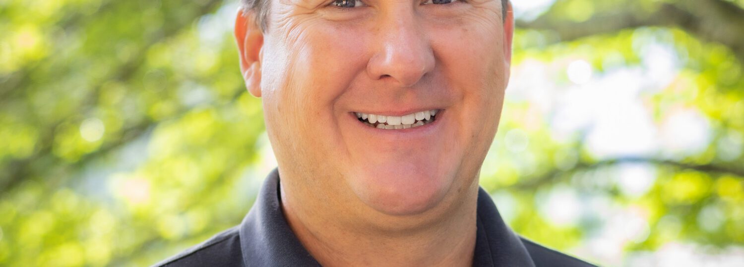 Headshot of Chacon wearing a black collar shirt in front of a natural background.