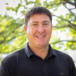 Headshot of Chacon wearing a black collar shirt in front of a natural background.