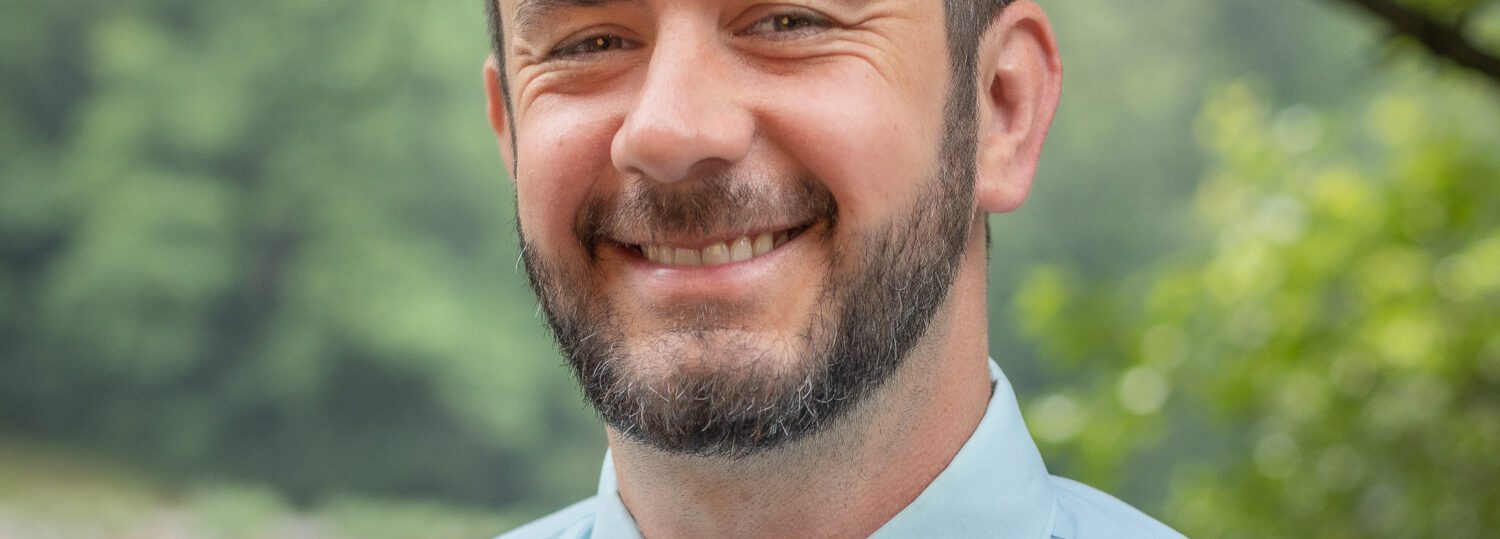 Headshot of Dillman with a light blue button up top and a blue and gray tie.