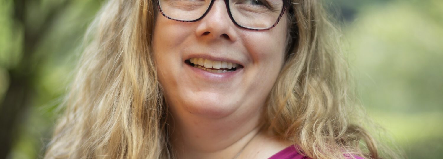 Headshot of Hawley wearing a dark pink blouse and standing in front of a natural backdrop.