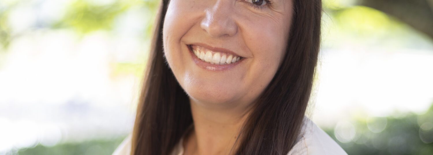 Headshot of Kelly wearing a white blouse before a natural background.