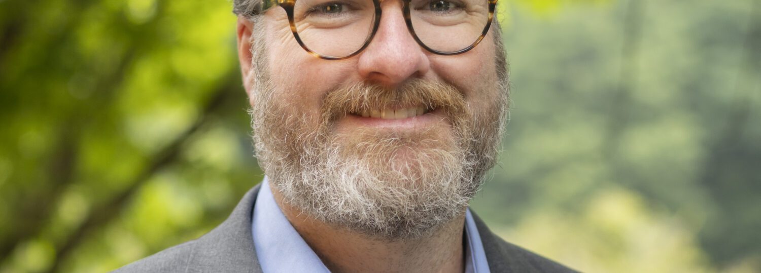 Headshot of Phelan wearing a gray blazer over a powder blue button up shirt.