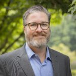 Headshot of Phelan wearing a gray blazer over a powder blue button up shirt.